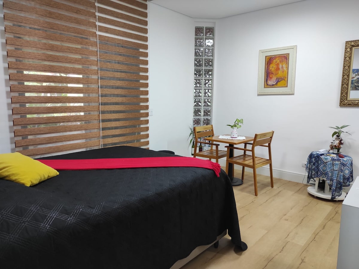 A spacious room features a large bed with a dark coverlet and a vibrant red accent. Natural light filters through wooden blinds. A small dining area includes a round table and two wooden chairs. Decorative plants and artwork add a touch of color to the setting.
