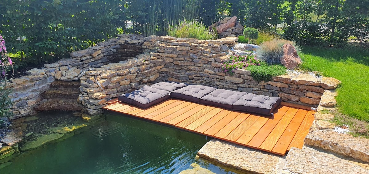 A wooden platform extends over the water, featuring comfortable seating pads. Natural stone elements form a gentle waterfall cascading into the pond, surrounded by lush greenery and thoughtfully arranged ornamental plants.