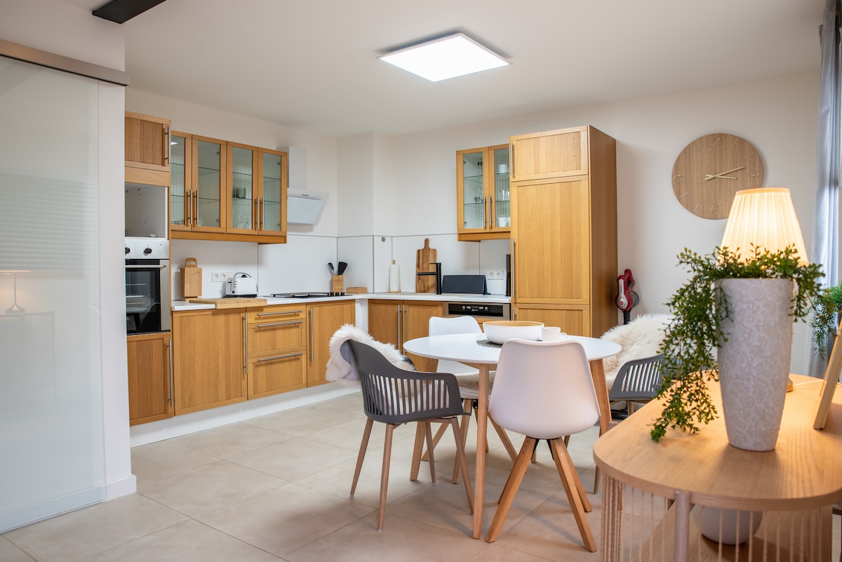 A modern kitchen is equipped with wooden cabinetry, stainless appliances, and ample countertop space. A round dining table with white and wooden chairs is positioned centrally, complemented by a decorative lamp and a potted plant. Light-colored tiles enhance the spacious feel.