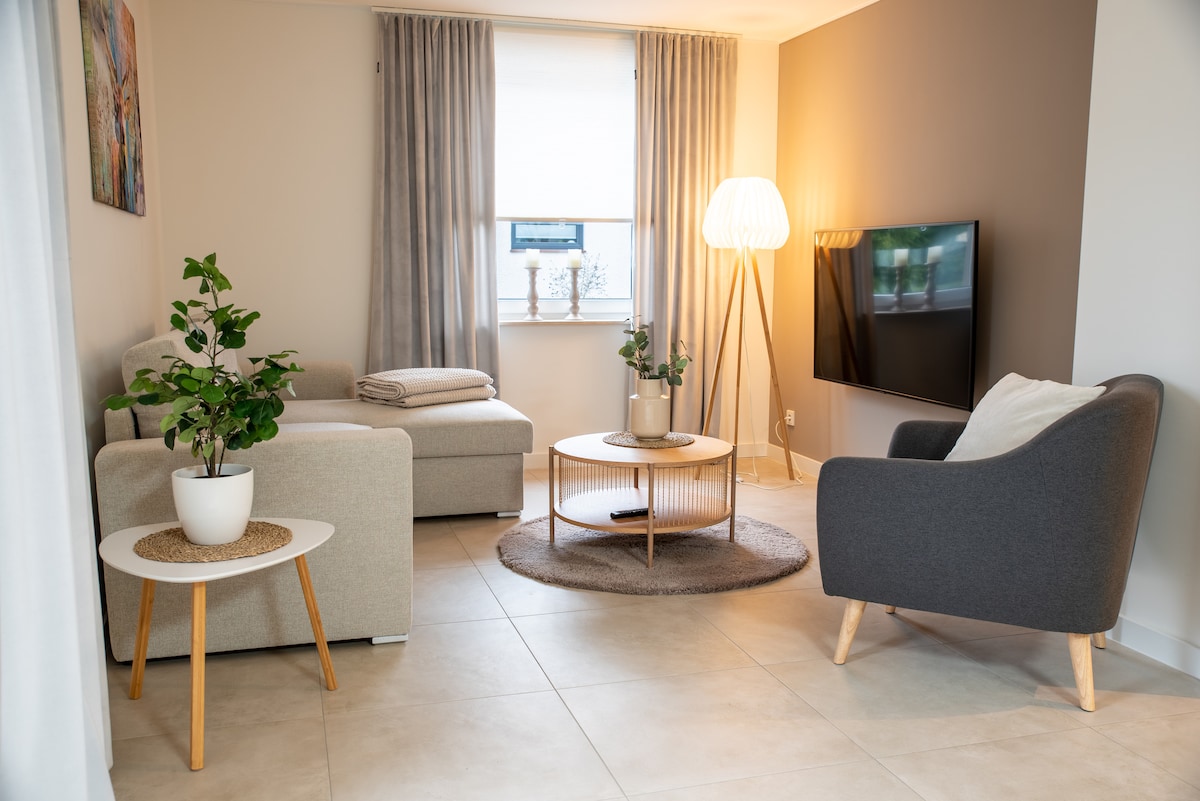 A modern living area is presented with a light grey sofa and a dark grey armchair, positioned around a round coffee table. Natural light enters through a window, complemented by soft curtains. A plant adds a touch of greenery, enhancing the inviting ambiance.