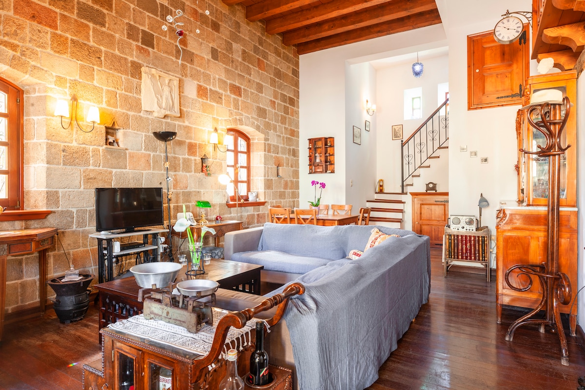 A spacious living area features a stone wall that adds texture to the room. A large, neutral-colored sofa is arranged around a dark wooden coffee table with decorative items. A TV is positioned against the wall, while a wooden dining set is visible in the background.