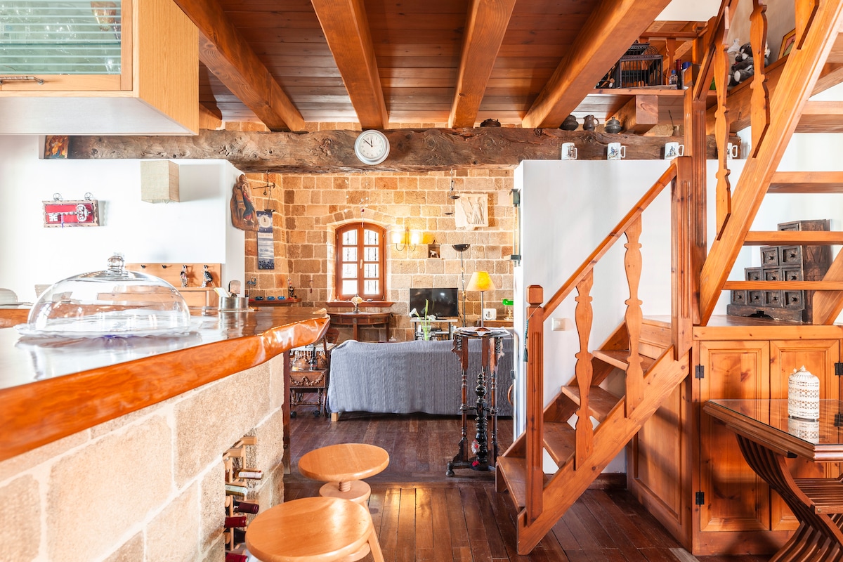 An open living space features wooden beams and a warm color palette. A rustic staircase leads to the upper level. A kitchen bar with stools provides a casual dining area, while a cozy seating arrangement with a soft grey blanket is visible, creating a welcoming environment.