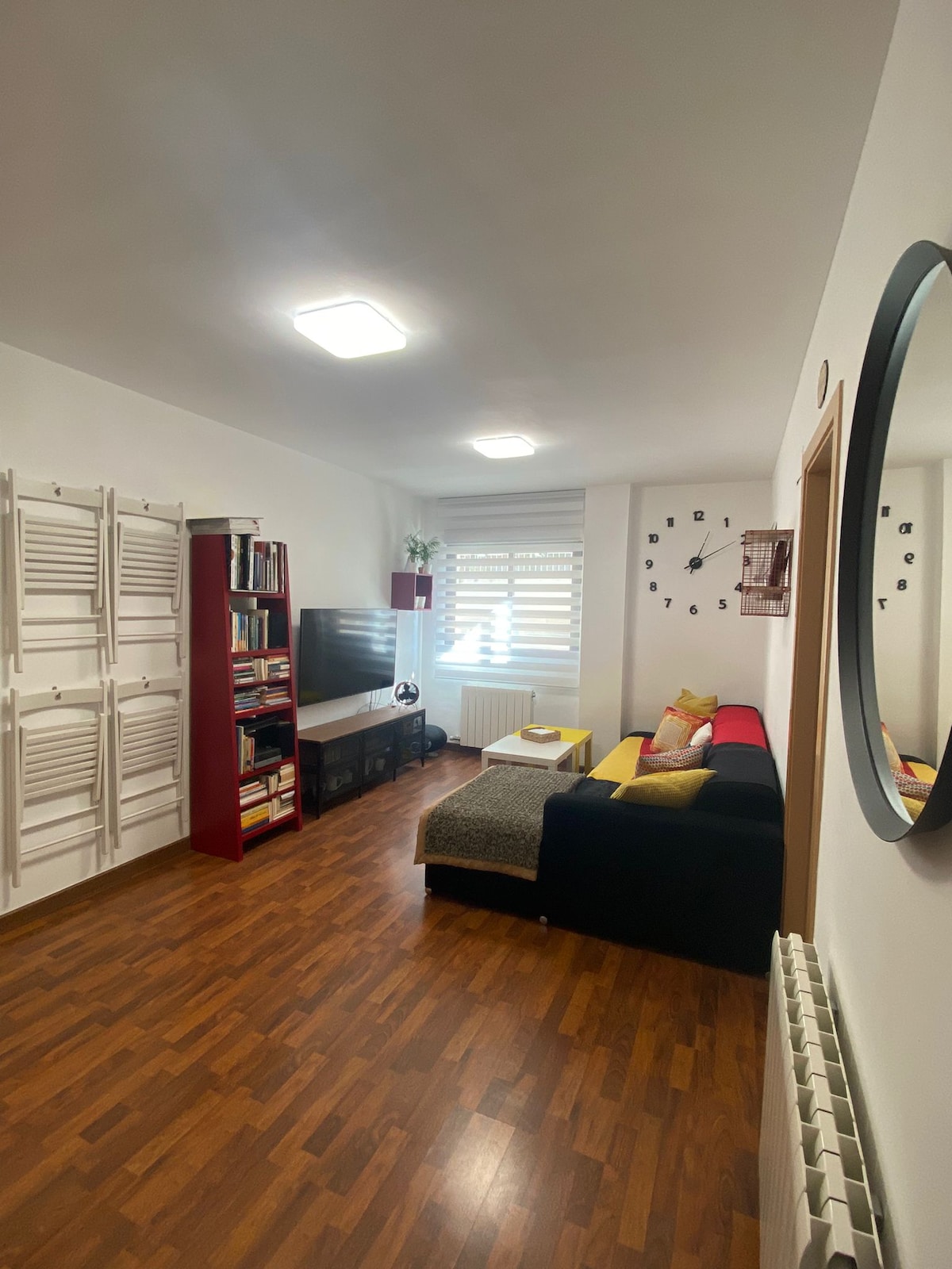 A cozy living area features a comfortable sofa adorned with decorative cushions, arranged next to a sleek media unit holding a television and books. Natural light filters through a window with horizontal blinds, highlighting the warm wood flooring and modern wall decor.