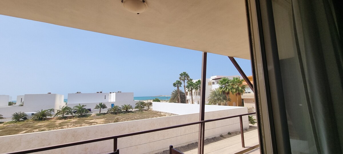 A view from the balcony reveals a stretch of the coastline and sea in the distance. The foreground features a landscape with palm trees and white buildings, enhancing the sense of openness and tranquility.