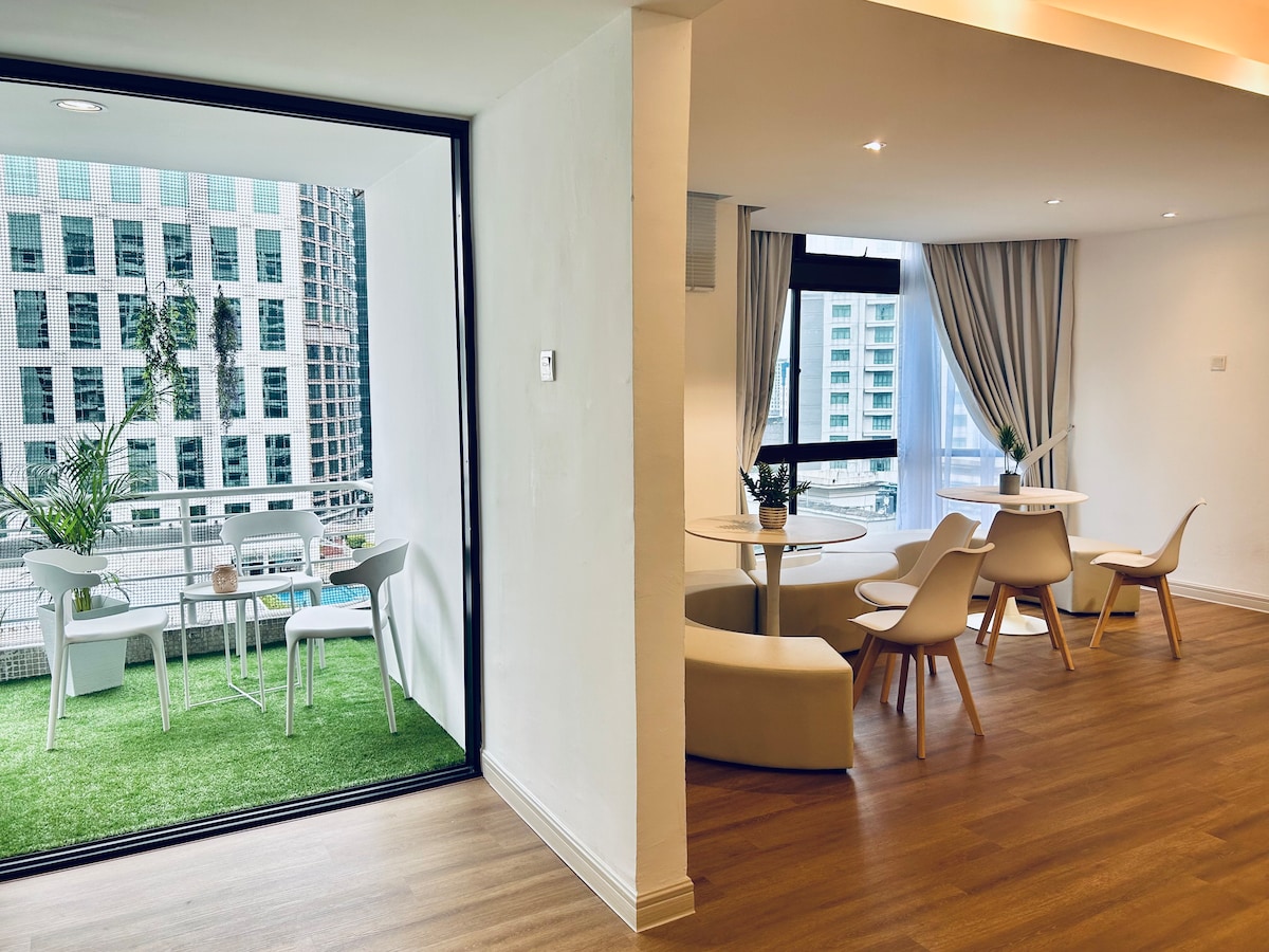 A bright and airy living space features a dining area with a round table and modern chairs. Adjacent is a balcony with green flooring, showcasing outdoor seating and greenery. Large windows allow natural light to illuminate the interior, offering views of the city skyline.