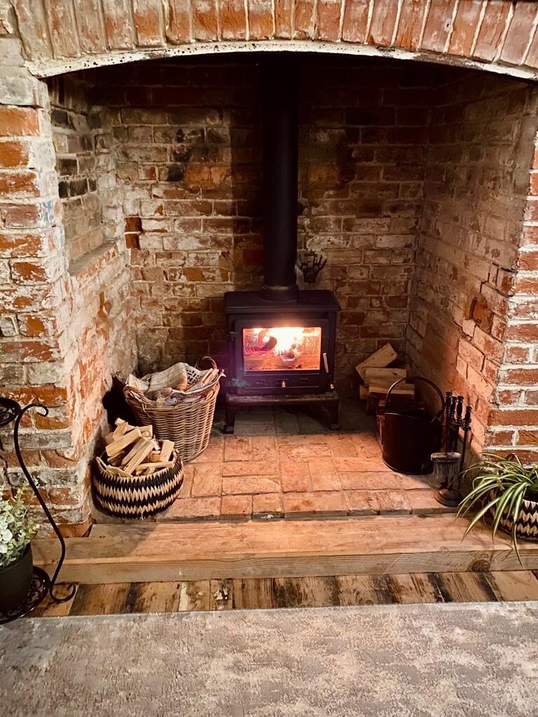 A charming wood-burning stove is nestled in a brick fireplace, emanating warmth. Nearby, a woven basket holds logs, while an additional metal container is present. The rustic brick wall complements the inviting atmosphere of the space.