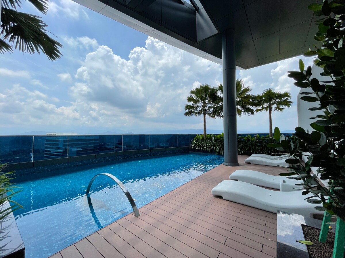 An outdoor infinity pool is surrounded by inviting loungers, offering sweeping views of the sky. Lush greenery and palm trees frame the area, providing a tranquil atmosphere. The pool's water reflects the clouds above, creating a serene environment for relaxation.