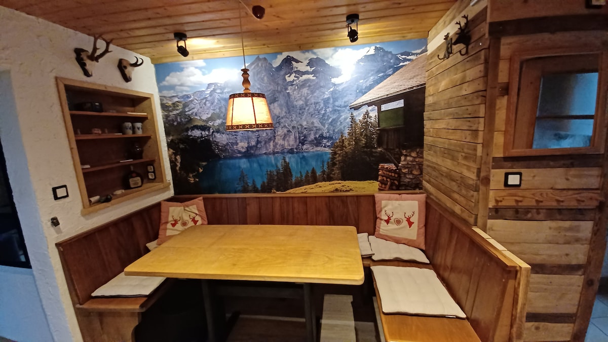 A wooden dining area features a large table surrounded by a cozy bench. Soft cushions add comfort, while a scenic mountain-themed mural serves as a backdrop. A pendant light hangs above, enhancing the rustic charm of the space.