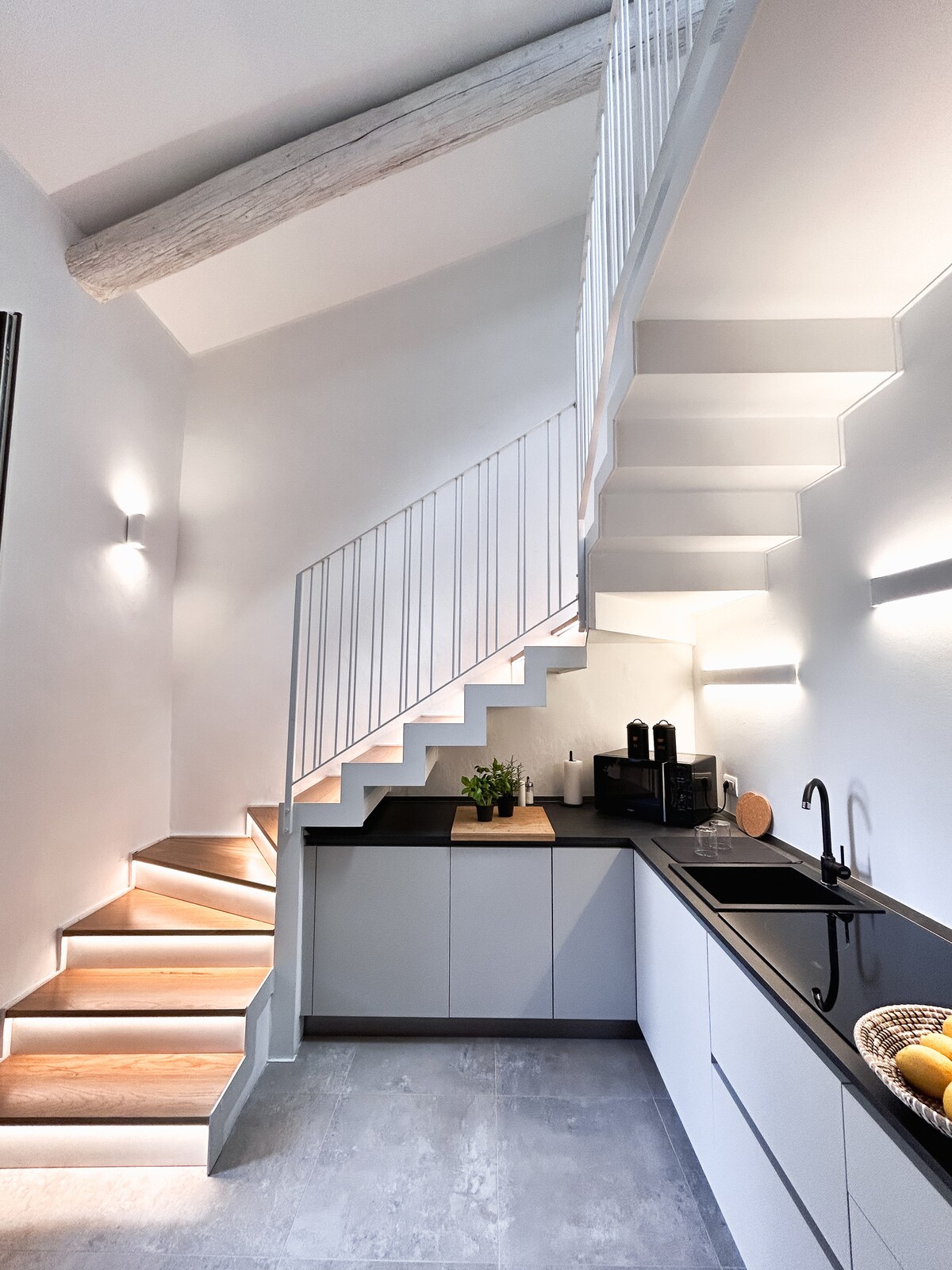 A modern kitchen is showcased with sleek cabinetry and a dark countertop. A staircase with illuminated steps leads to the upper level, enhancing the open design. Subtle lighting accents the wall, creating a bright and inviting cooking space.