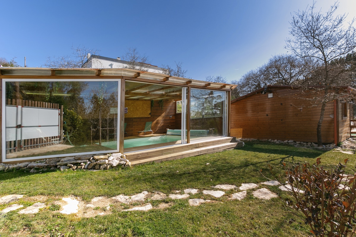 A wooden house surrounded by nature features large glass windows, showcasing an inviting indoor swimming pool. The well-maintained garden includes stone pathways and trees, providing a serene outdoor environment. A secondary wooden structure stands nearby, highlighting the rustic charm of the property.