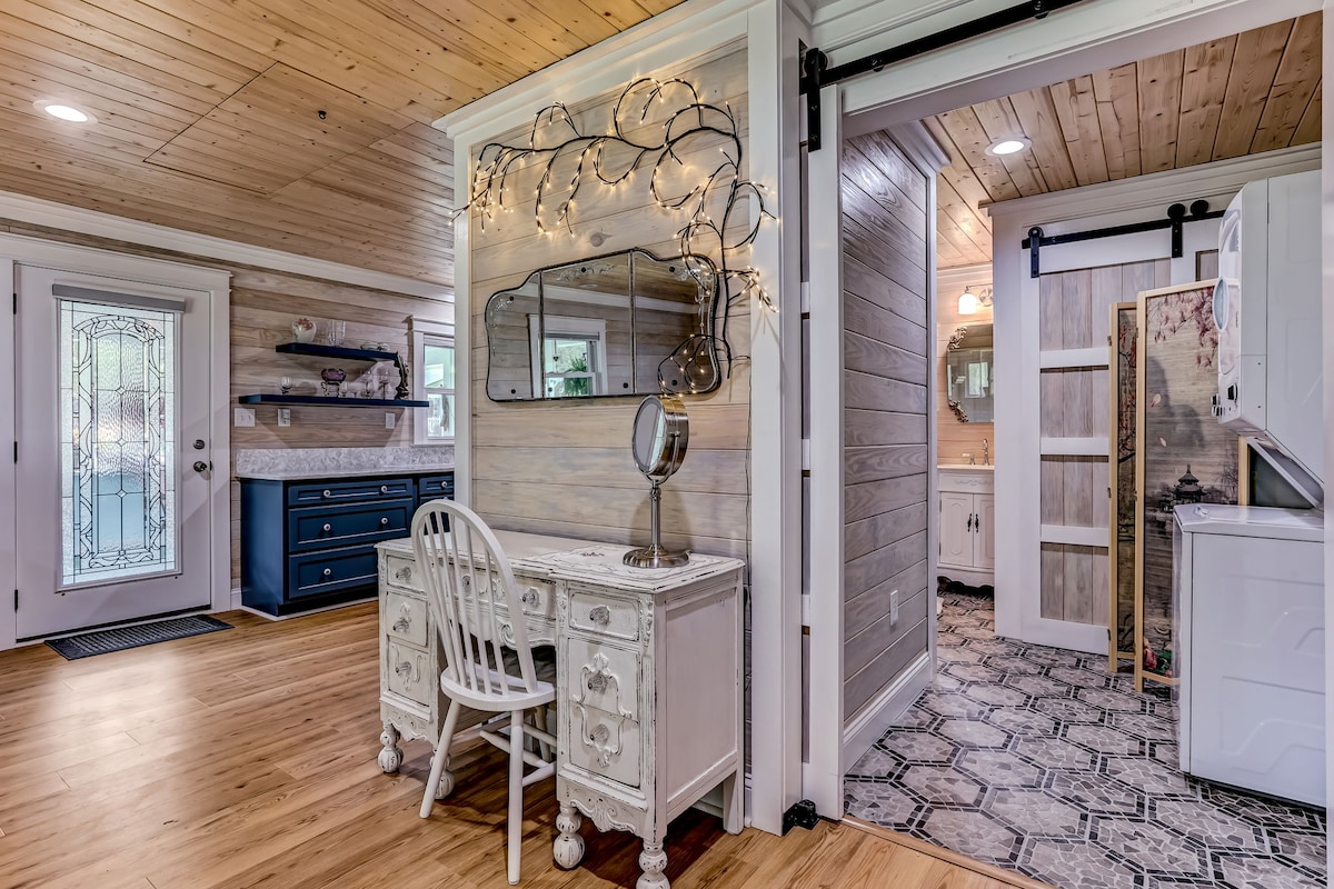 The entrance area features warm wooden paneling and a decorative mirror illuminated by soft lighting. A white desk with a vintage design is positioned beside a blue dresser. The doorway leads to a bathroom, visible through a sliding barn door.