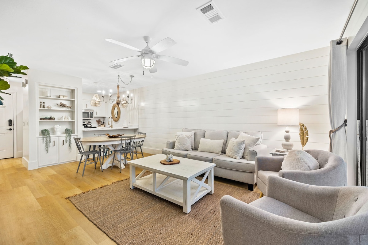 A bright living area is showcased with natural wood flooring and warm lighting. Comfortable seating is arranged around a spacious coffee table, complemented by a textured area rug. A dining area with light chairs is visible in the background, enhanced by a decorative light fixture.