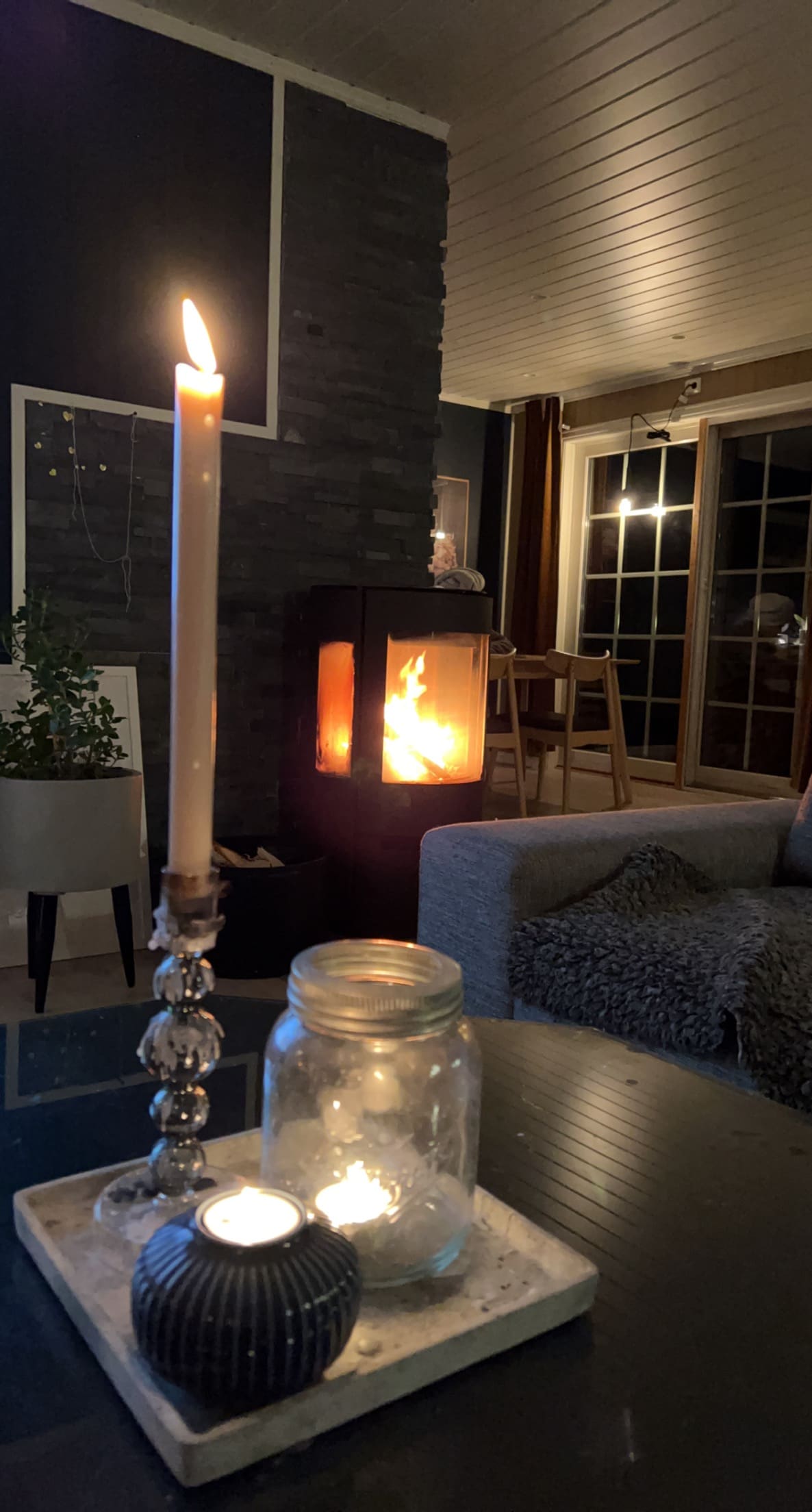 A warm living space is featured, showcasing a lit fireplace and candle arrangement on a low table. A comfortable sofa is positioned nearby, and large windows provide views of the outdoor area. Soft lighting enhances the cozy ambiance.