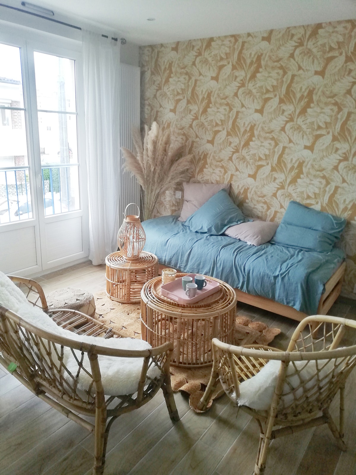 A cozy seating area features a light-colored wooden sofa with soft blue cushions, surrounded by two rattan armchairs and a small round table. Natural decor elements, including dried plants, complement the leafy wallpaper, while sunlight highlights the space through large windows.