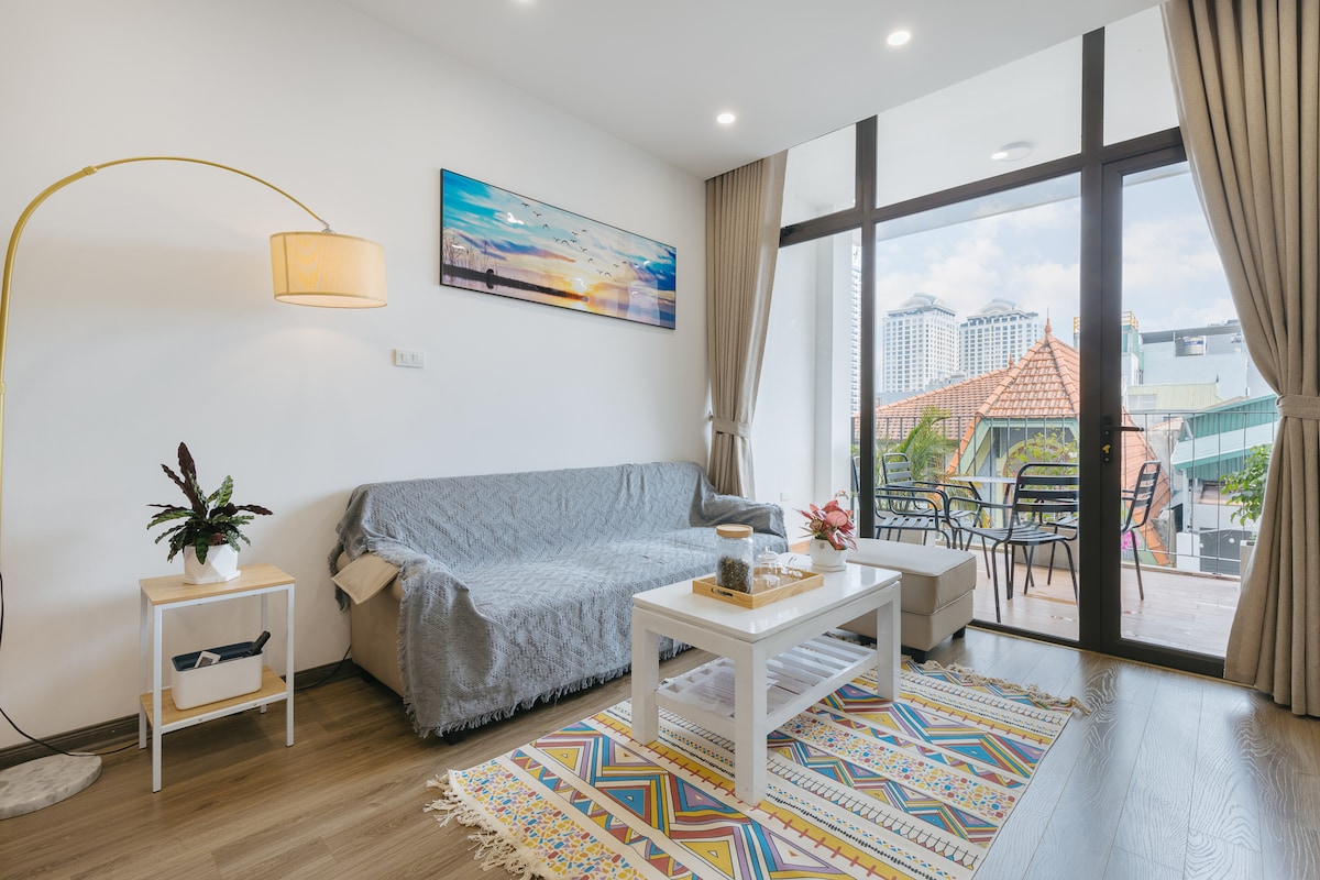 A modern living space is showcased with a light gray couch draped with a soft cover. A coffee table sits atop a colorful area rug, with a lamp illuminating the room. Large glass doors lead to a bright balcony with rooftop views.