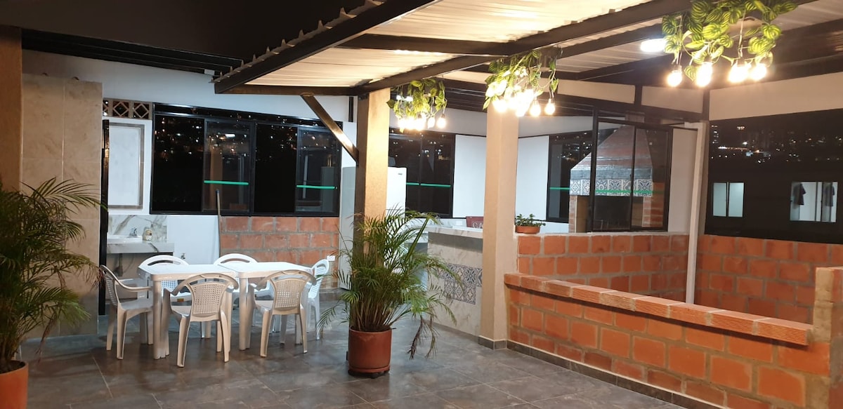 An outdoor terrace area is seen, featuring a large open space with a decorative ceiling and hanging lights. A round table surrounded by several chairs is positioned in front of a semi-enclosed dining or social area. Green plants add a touch of nature to the space.