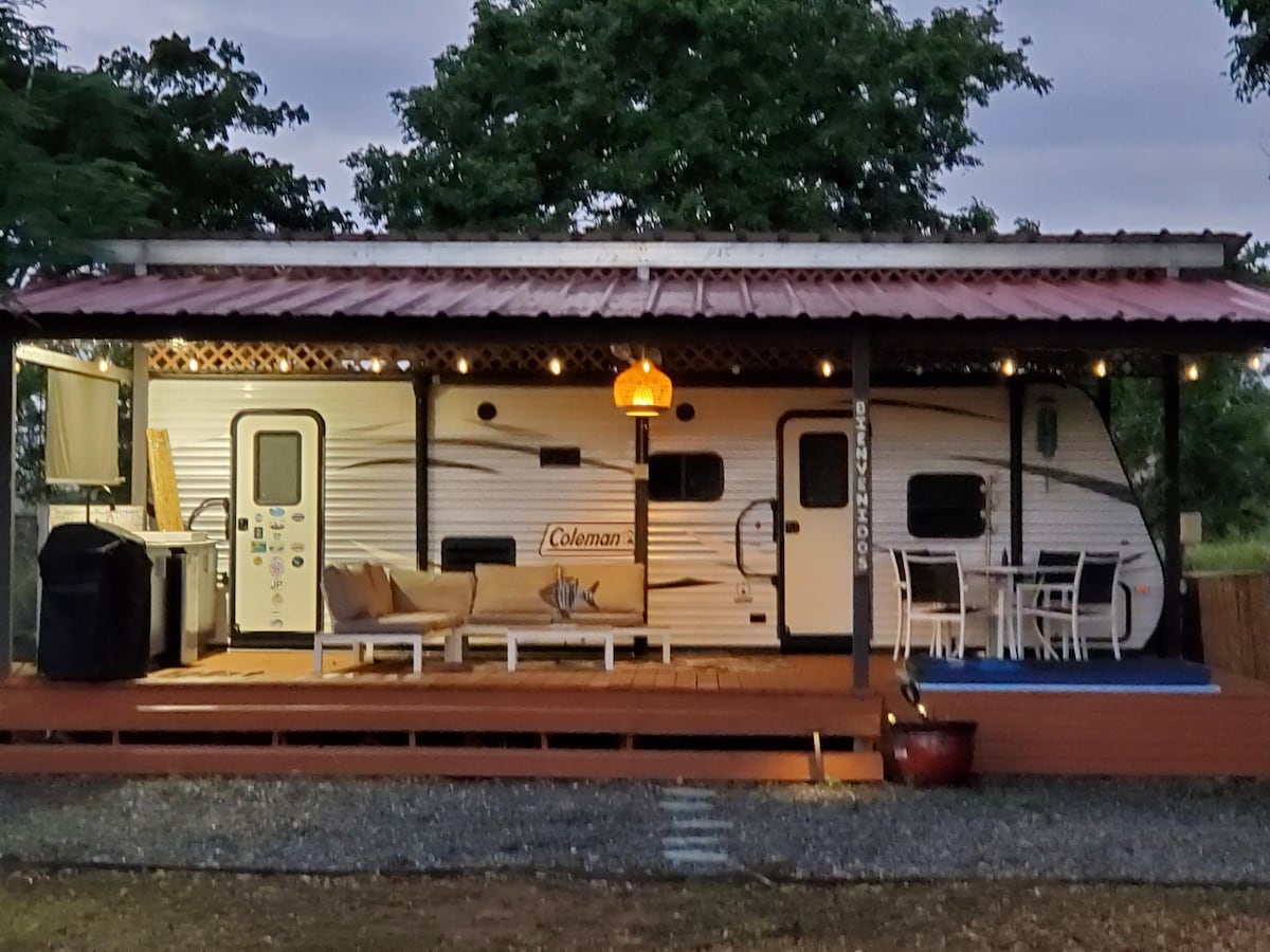 A camper is nestled under a covered patio, adorned with string lights that create a warm glow. An outdoor kitchen, featuring a sink and grill, is positioned to the left. A cozy seating area with a sofa and dining table is situated on the patio, surrounded by lush greenery.