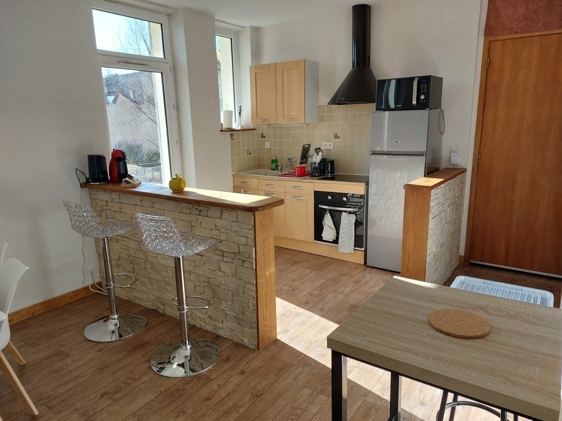 A modern kitchen is displayed, featuring light wood cabinetry and stone accents. Bar stools are positioned at a counter, while a stainless steel refrigerator and stove are visible. Natural light floods the space through large windows, enhancing the warm tones of the wooden flooring.