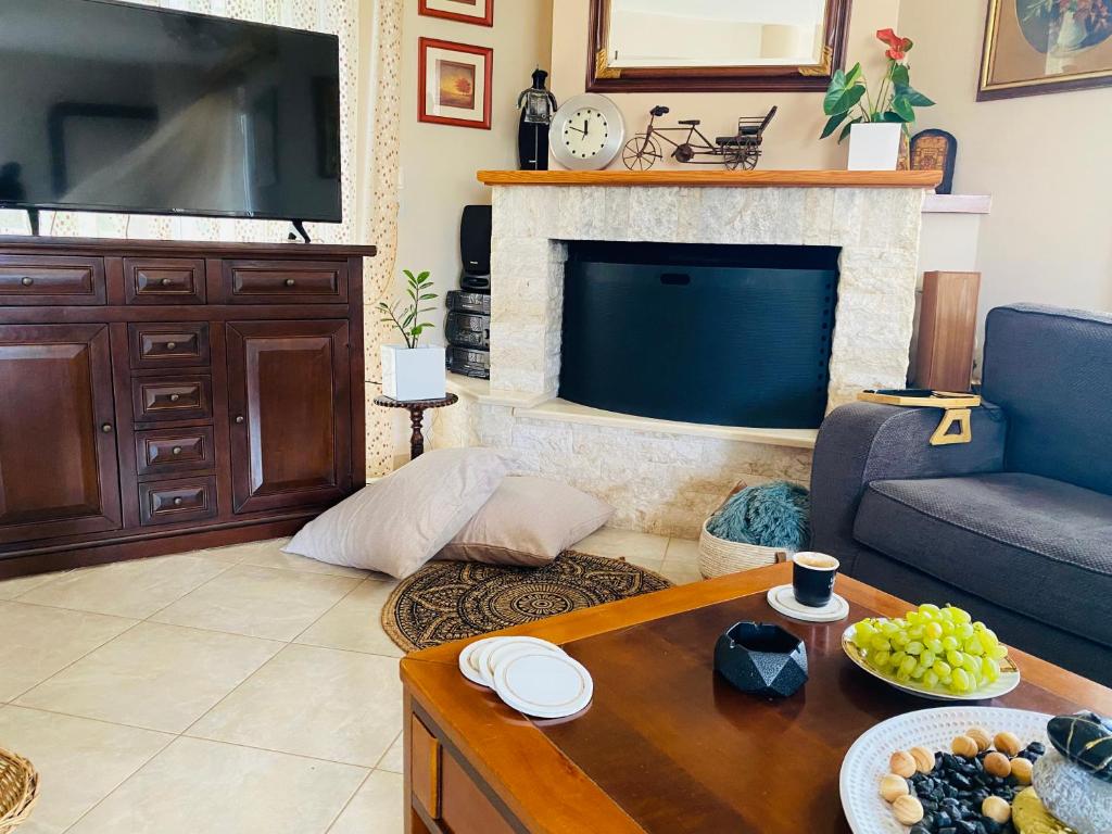 A living area is presented with a cozy sofa and a wooden coffee table. A flat-screen TV is placed on a cabinet, which contrasts with a stone fireplace. A decorative bowl of grapes and snacks is arranged on the table, accompanied by white plates and a cup of coffee.