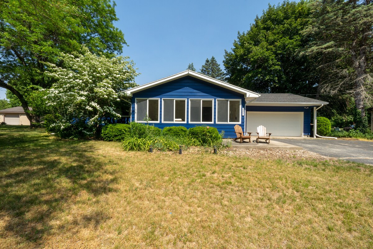 A blue exterior of a single-story cottage is presented, surrounded by lush greenery. Large windows allow natural light to fill the living space. Two wooden adirondack chairs are positioned on the front lawn, inviting relaxation in a tranquil setting.