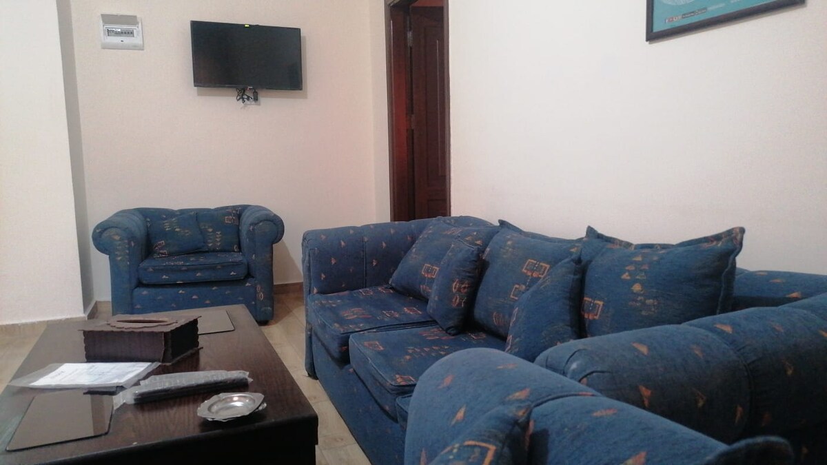A comfortable living area is featured, furnished with a blue fabric sofa and armchair. A coffee table is positioned in front of the seating, and a wall-mounted television is visible, contributing to a relaxed environment. Natural light illuminates the space.