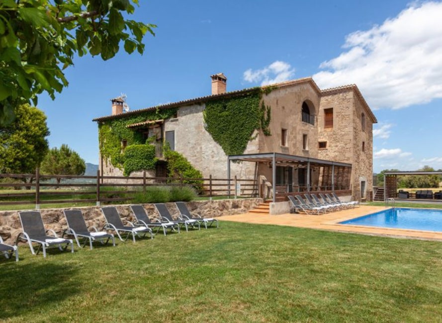 The exterior of the villa displays a rustic stone facade, partially covered with greenery. A spacious lawn leads to an inviting outdoor swimming pool, surrounded by lounge chairs. The patio area is covered, providing a shaded space for relaxation and gatherings.