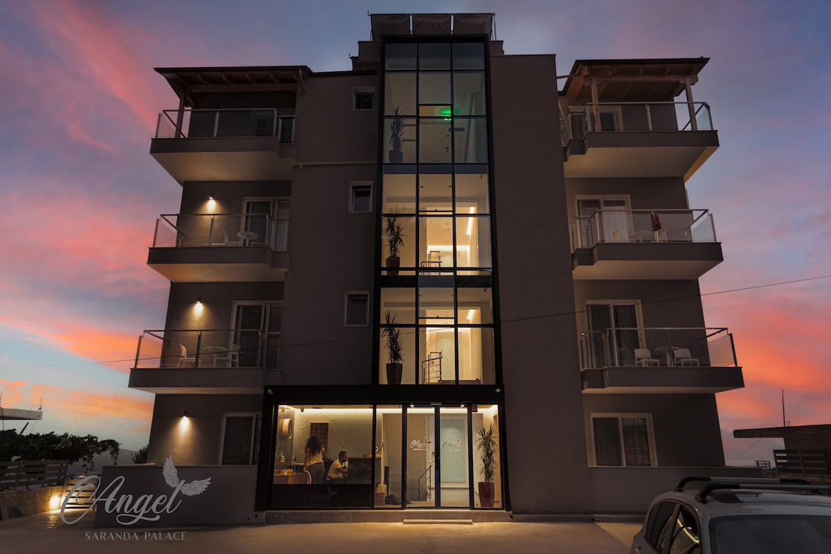 A modern multi-story building features large glass panels, showcasing its contemporary architecture. Balconies with railings extend from the upper floors, providing outdoor space. The entrance is highlighted by soft lighting, creating a welcoming atmosphere as twilight settles in the background.