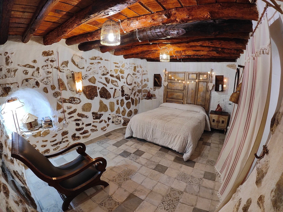 A rustic bedroom features a spacious bed with a textured white coverlet. Exposed wooden beams highlight the ceiling, while natural stone walls add character. Soft lighting is provided by hanging fixtures and wall sconces, enhancing the calm atmosphere. A comfortable chair is situated beside the bed.
