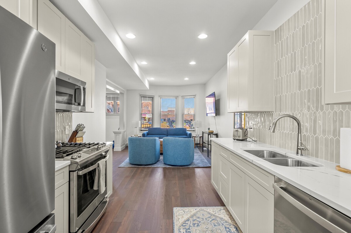 A contemporary kitchen is featured with stainless steel appliances, including a gas stove and refrigerator. The open layout connects to a bright living area, showcasing two blue sofas and large windows that provide natural light and views of the surroundings.