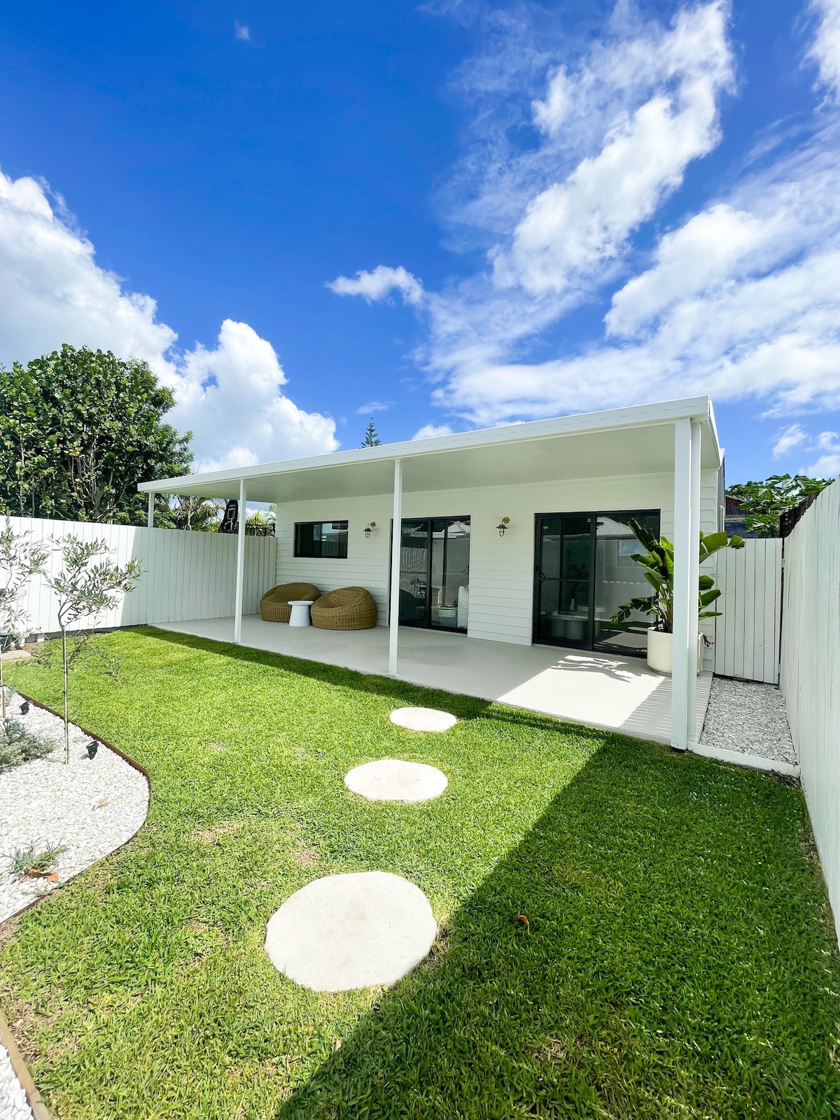 A spacious outdoor area is depicted, featuring a covered patio that provides shade. The garden showcases well-maintained grass and a pathway made of circular stepping stones. Lush green plants are arranged against the white fencing, under a clear blue sky dotted with a few clouds.
