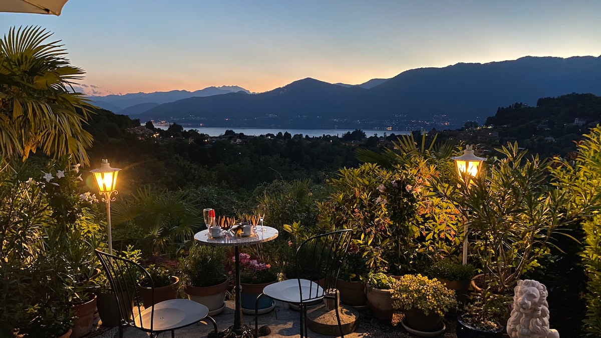 A serene outdoor terrace is surrounded by lush greenery, showcasing a small round table with two black chairs. Soft lighting from two lamps creates a welcoming ambiance while stunning views of mountains and the lake are visible at dusk.