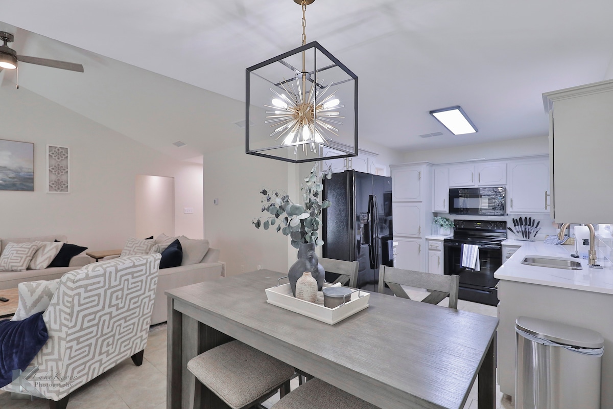 A modern dining area is featured with a rectangular table set for four. Above, a geometric light fixture provides illumination. The kitchen is visible, showcasing sleek black appliances and a bright countertop. A cozy living area with light-colored sofas is positioned to the left.