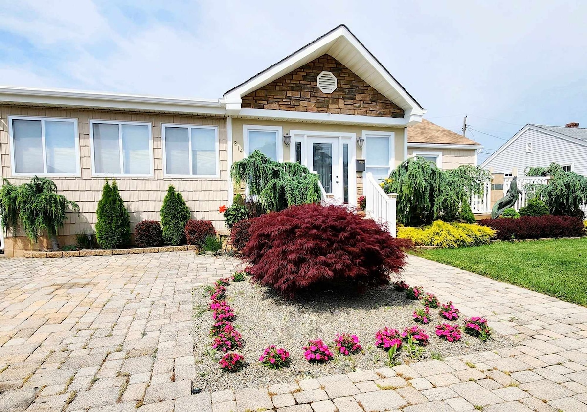 The exterior of the home features a landscaped yard with various shrubs and flowerbeds. A stone accent highlights the entrance, framed by large windows. Paved pathways lead to the inviting front door, surrounded by colorful blooms and lush greenery.