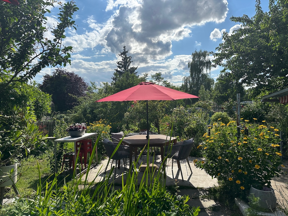 A spacious outdoor dining area is shaded by a large red umbrella, surrounded by lush greenery and colorful flower beds. A wooden table is accompanied by several chairs, offering a peaceful setting for meals and gatherings.