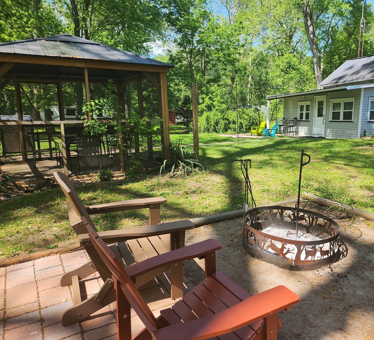 An outdoor space features a screened gazebo with dining chairs and a table, surrounded by lush greenery. Two wooden Adirondack chairs are placed near a fire pit on a stone patio, offering a relaxing spot to unwind in nature.