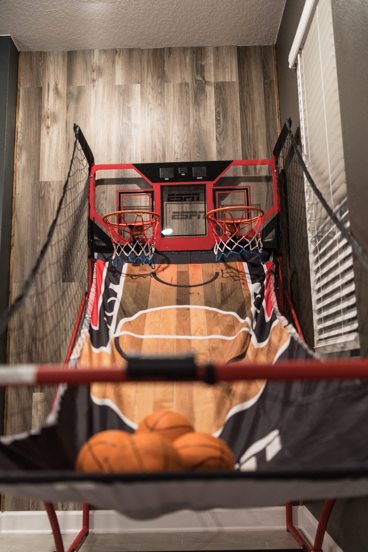 An indoor basketball arcade game is presented, showcasing two hoops set against a wooden backdrop. Bowling balls sit on the floor, inviting play. The design features a basketball court motif, with scoring electronics and regulation-style nets. Natural light filters through nearby window blinds.