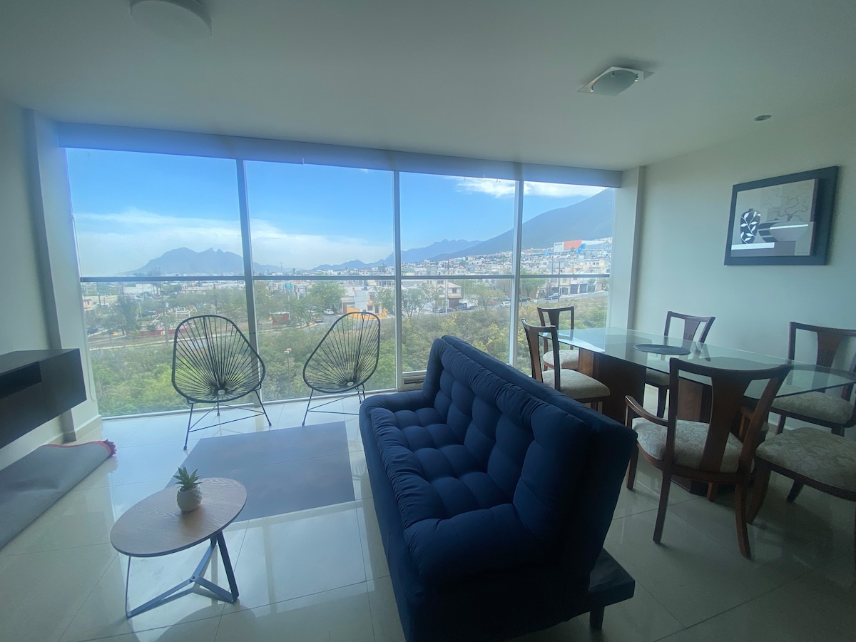 A modern living area features a blue sofa paired with a circular coffee table. Two wire-frame chairs are positioned nearby, offering a view through large windows of the surrounding Monterrey landscape and mountains in the distance. A dining table with chairs is also visible.