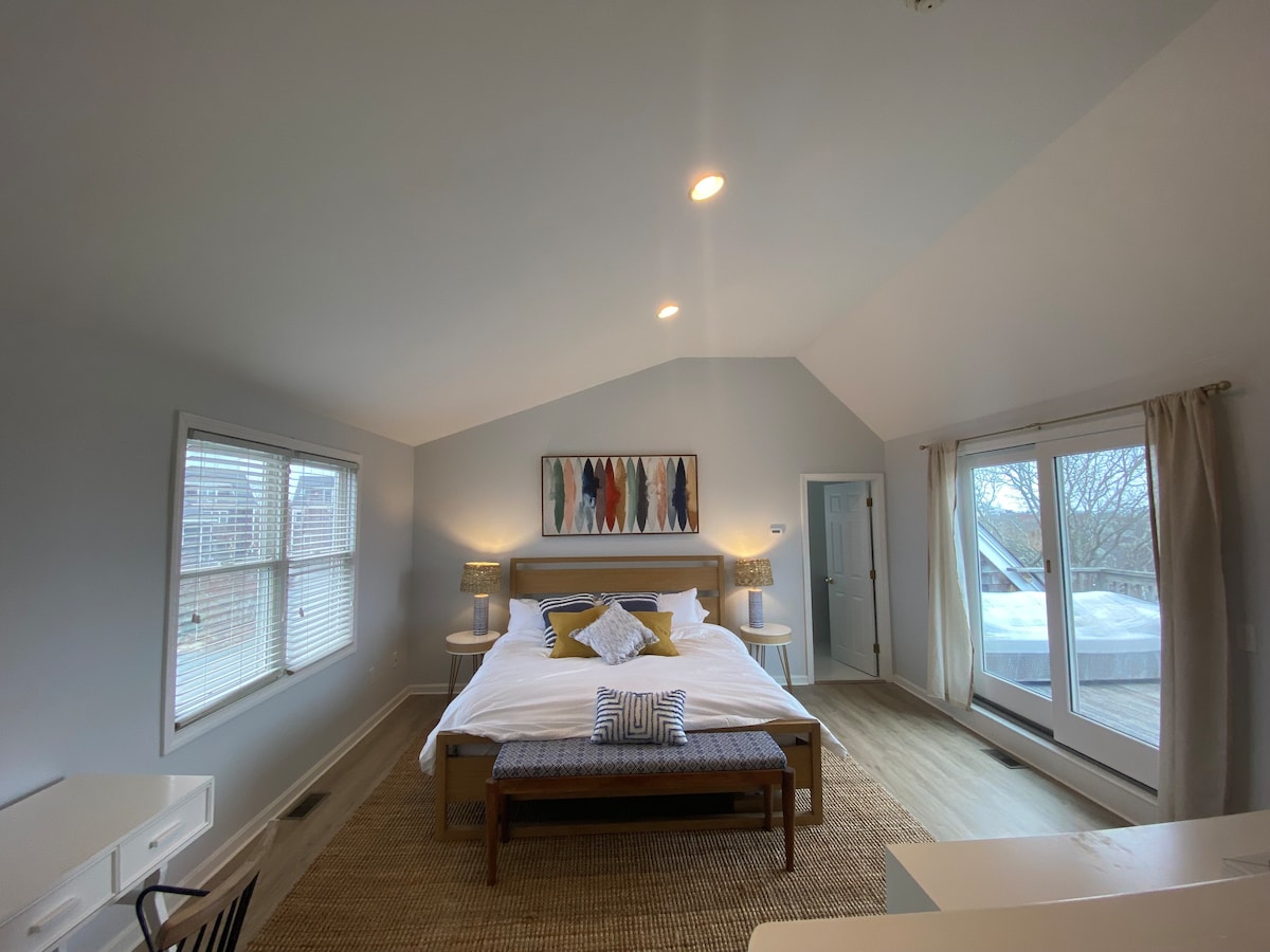 A spacious bedroom features a large bed with white linens and decorative pillows. Light filters through multiple windows, highlighting the soft gray walls. A small writing desk is positioned nearby, and a bench is placed at the foot of the bed, creating a functional and comfortable environment.