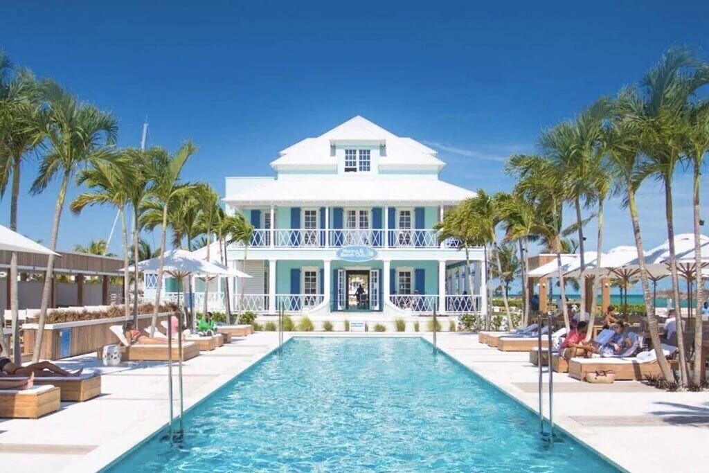 A spacious outdoor pool is framed by lounge chairs and umbrellas, leading to a grand building with light blue exterior. Palm trees provide shade, and clear skies create a bright atmosphere. The pool reflects the vibrant surroundings, inviting relaxation.