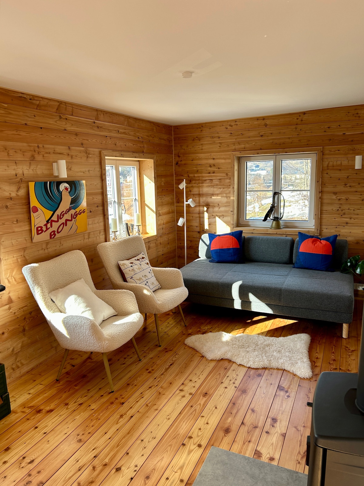 A cozy living area features a gray sofa and two light-colored armchairs with wooden legs. Natural wood walls and a soft rug enhance the inviting ambiance. Two windows allow light to enter, and a small lamp stands beside the sofa, contributing to a warm and comfortable setting.