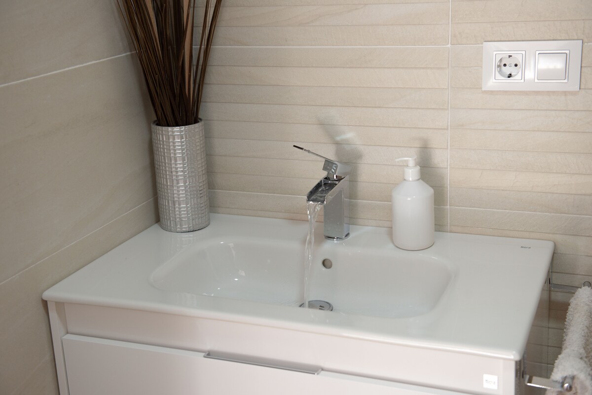 A modern bathroom sink area is displayed, featuring a sleek white countertop with an integrated basin. Water flows from a polished chrome faucet, complemented by a stylish soap dispenser and a metallic vase containing decorative branches.