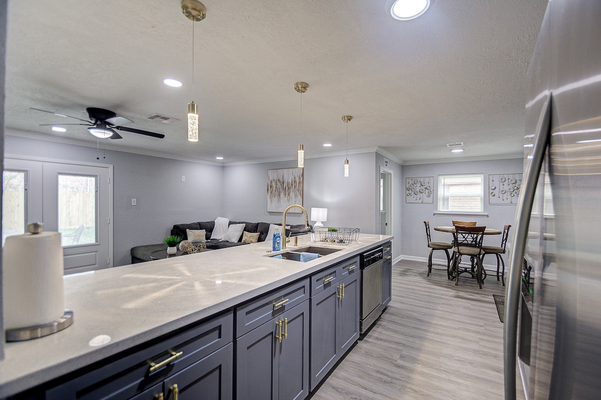 A spacious kitchen area features modern cabinetry and a large island with a sink. An open layout connects to a cozy living area with a sectional couch. A dining table is positioned nearby, while natural light filters through several windows.