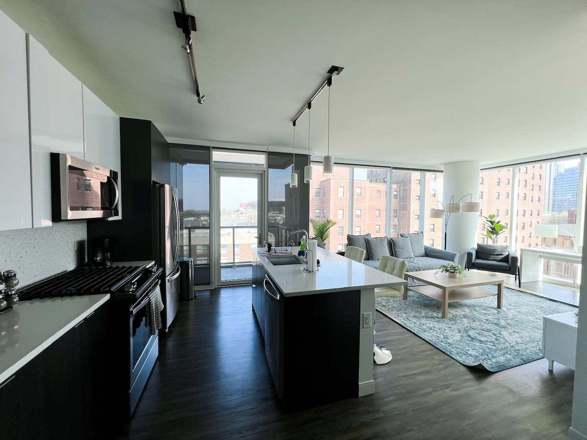 A modern, open-concept kitchen and living area showcases sleek white cabinetry and stainless steel appliances. A spacious island with seating separates the kitchen from a cozy sitting area, featuring a large sectional sofa and a coffee table. Natural light floods the space through expansive windows.