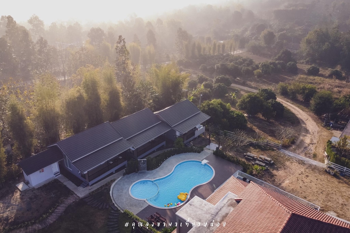 An aerial view captures a serene landscape, highlighting a curved swimming pool surrounded by lush greenery. The property features modern dwellings with dark roofs set against the backdrop of rolling hills and a winding path leading through the trees.