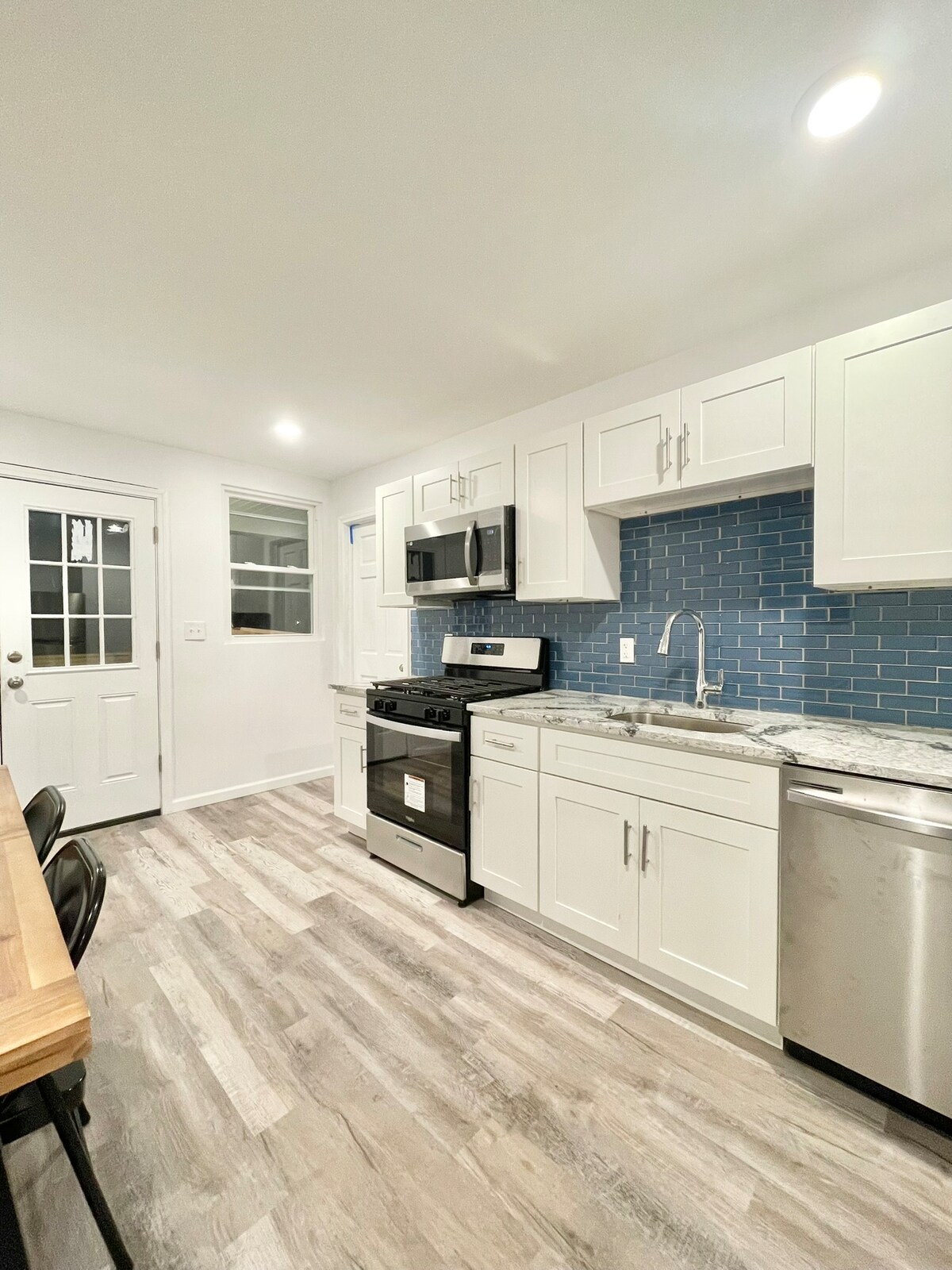A modern kitchen is presented with white cabinetry and granite countertops. Stainless steel appliances, including a stove, microwave, and dishwasher, complement blue tiled backsplash. A door leads to the exterior, while a wooden dining table with black chairs creates a functional dining space.
