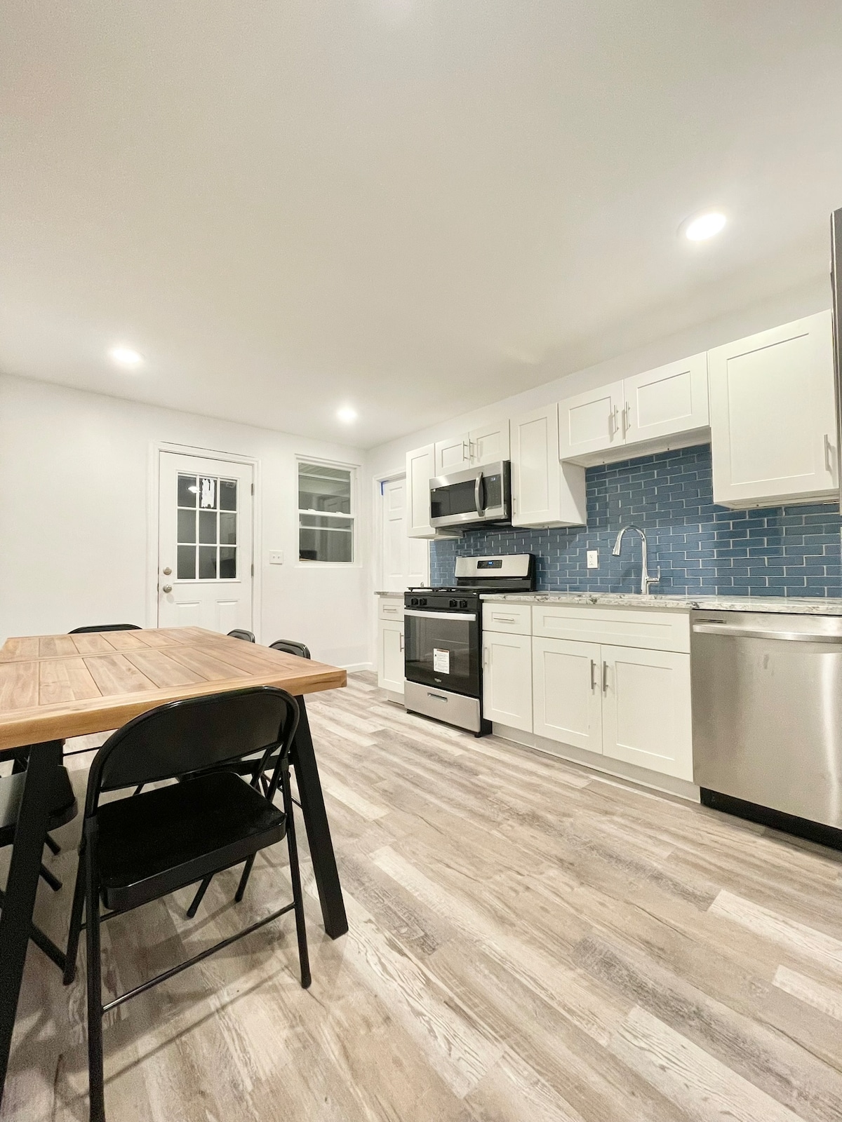A modern kitchen is featured with white cabinetry and stainless steel appliances. A blue tiled backsplash complements the spacious countertops. A wooden dining table with black chairs is positioned in the foreground, while a door to the outside provides additional light.
