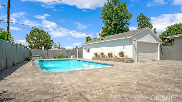 A spacious backyard features a well-maintained pool surrounded by a large patio area. Lush greenery and trees are visible in the background, providing a natural setting. A single-story structure, with a garage, is situated adjacent to the pool, contributing to the overall openness of the space.