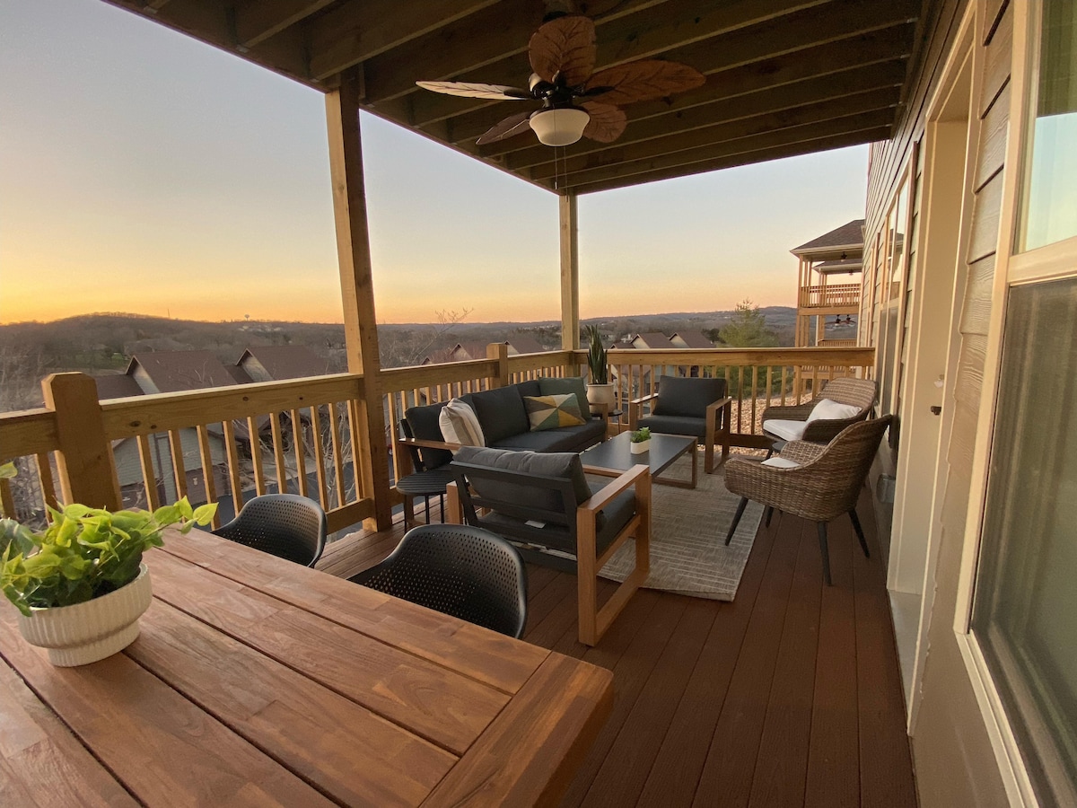 An outdoor deck is furnished with a seating area that includes a couch and chairs, offering a space for relaxation. A dining table is also present, complemented by the backdrop of a scenic sunset over distant hills.