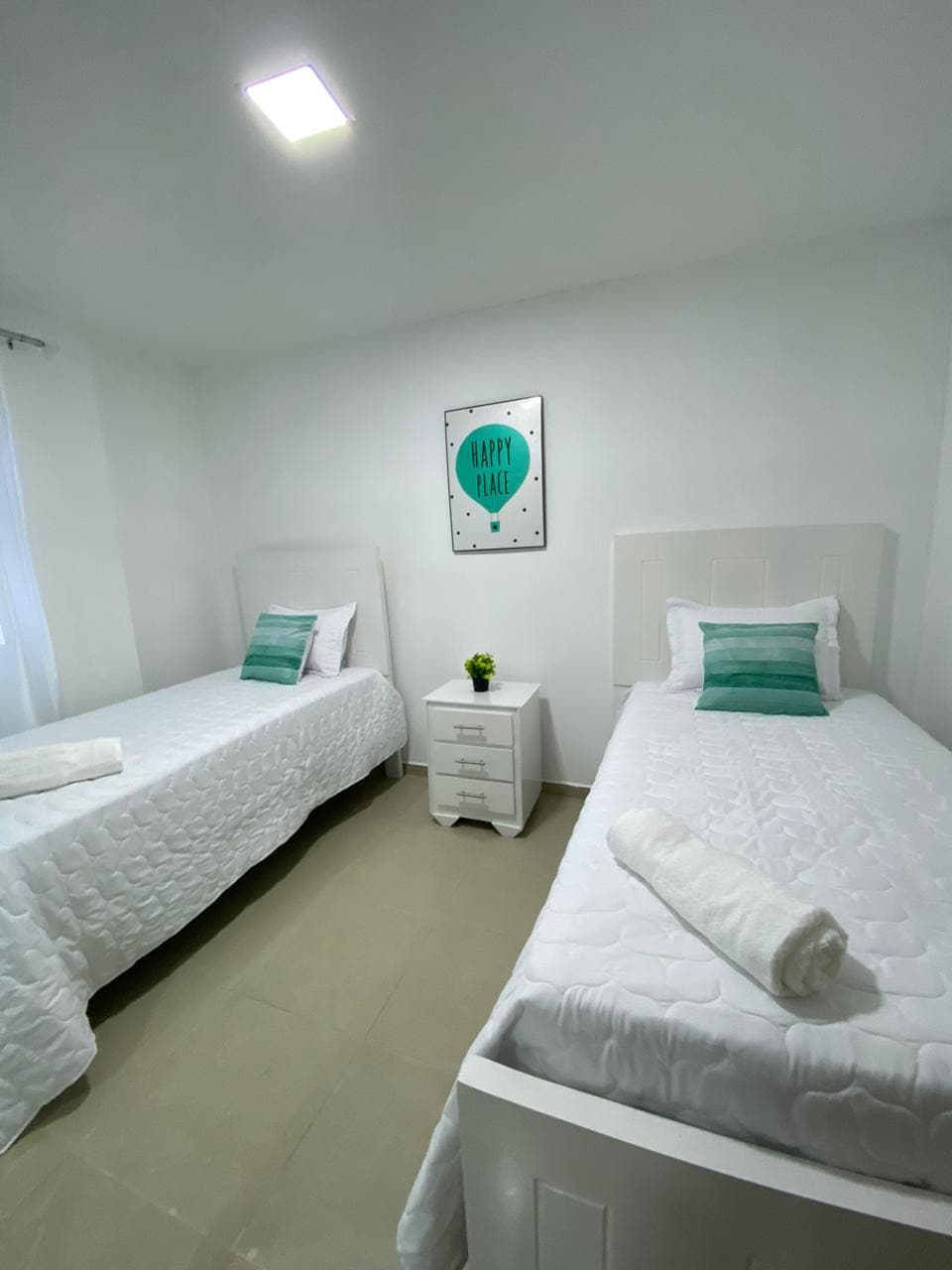 The room features two twin beds with white bedding, accented by turquoise decorative pillows. A small bedside table is positioned between them, topped with a potted plant. Natural light enters through a nearby window, complemented by a cheerful wall decoration.