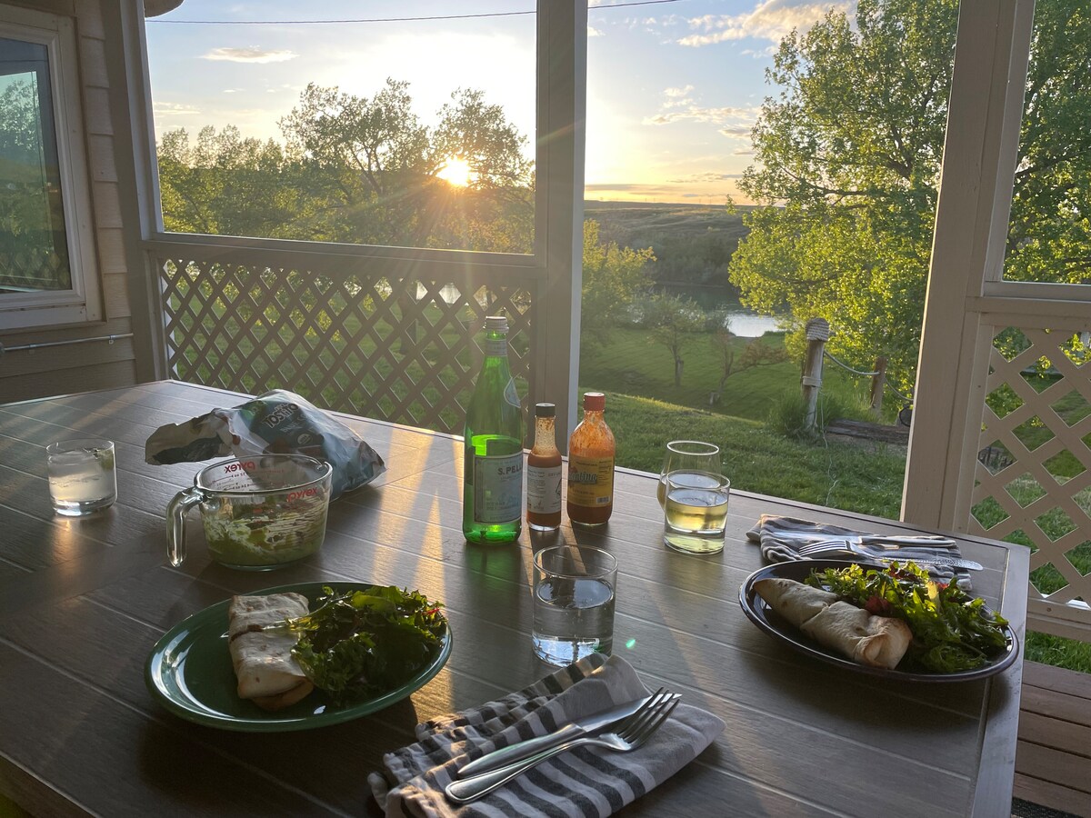 A dining table is set with plates of food, accompanied by drinks and condiments. The scene is enhanced by the sight of a sunset through large windows, with greenery visible outside and the North Platte River in the background.